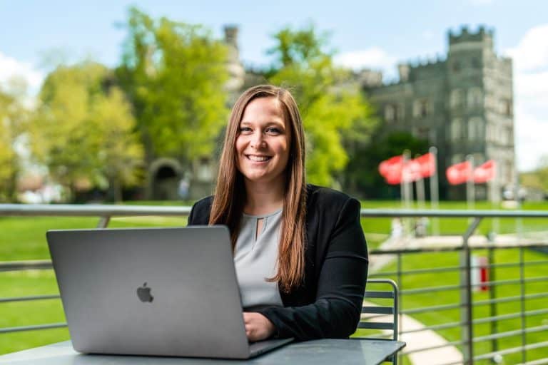 A woman smiling at the camera while on her laptop at a table outdoors in front of Arcadia University